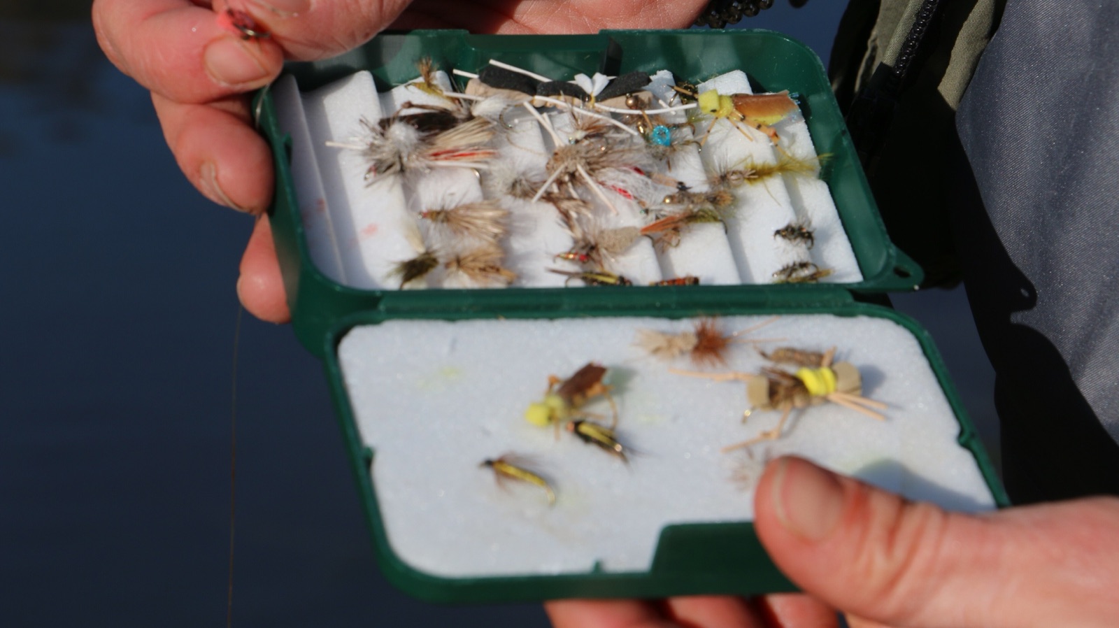 A fly-fishing box of tied flies held over water.